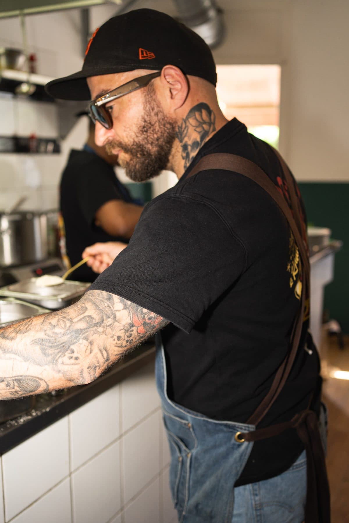Alessandro at the pass: cap, sunglasses, tattooed arms, denim apron
