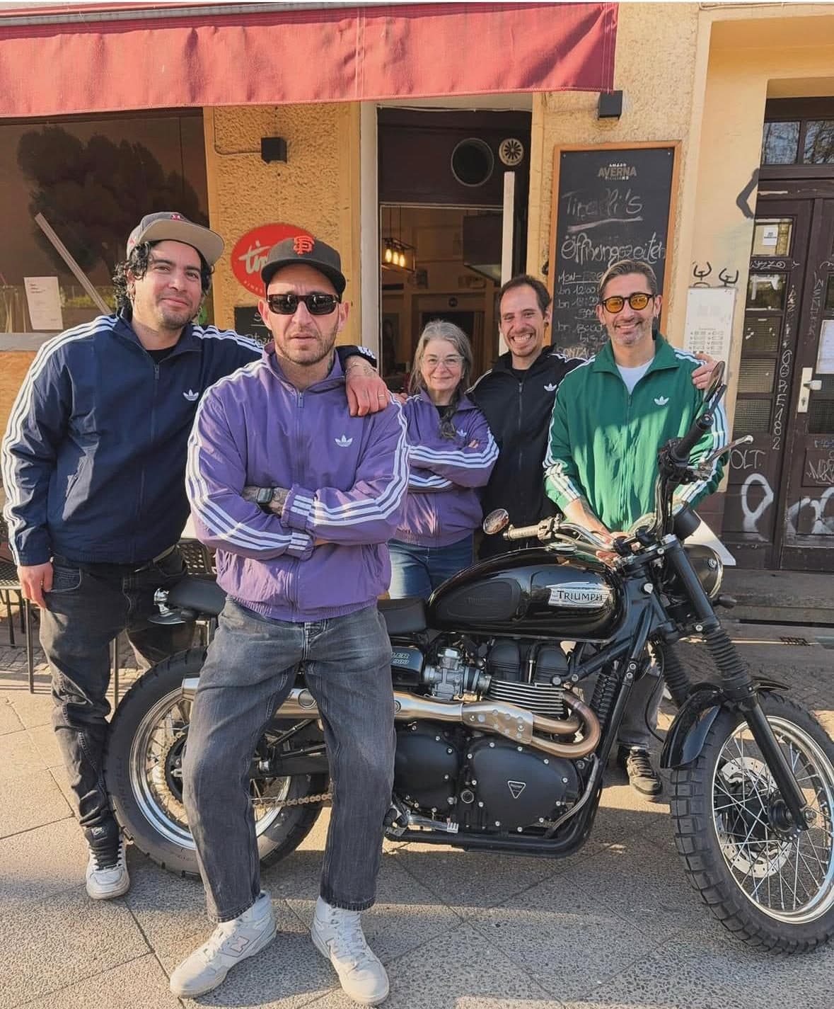 Die Squadra von Tirelli's in Trainingsjacken vor dem Laden, vorne eine schwarze Triumph